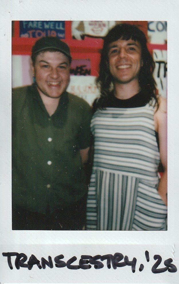 Two people are smiling in front of colorful signs, with "Transcestry, '28".