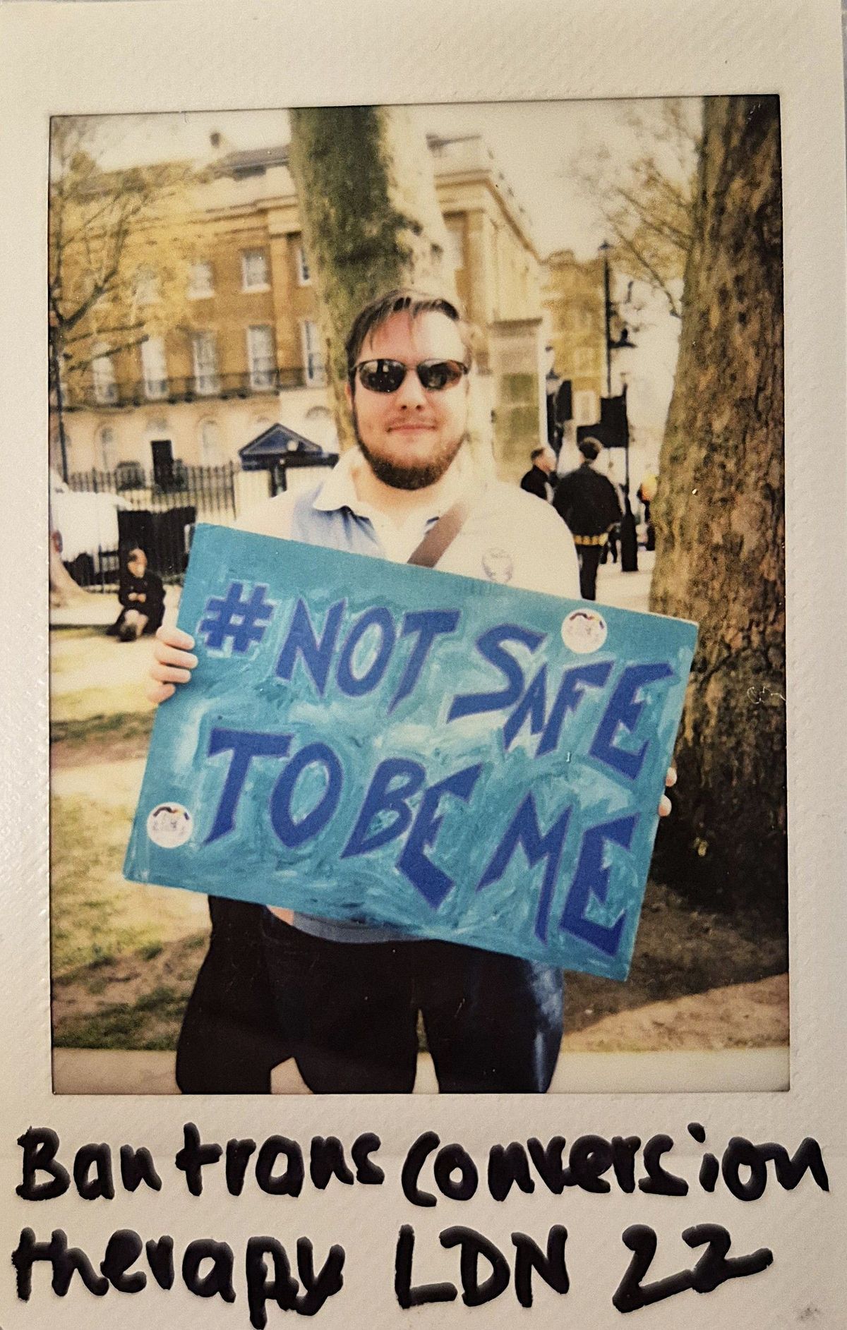 A person holding a sign that reads "#NOT SAFE TO BE ME" stands outside, wearing sunglasses.