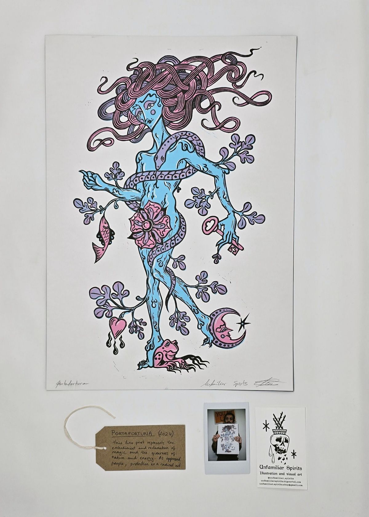 A print, entitled "PORTAFORTUNA", of a plant-like figure with intricate details and flowing hair. Colored in blue and pink on a white background. With a brown, handwritten luggage tag, a Polaroid of the artist and a contact card for 'Unfamiliar Spirits'.