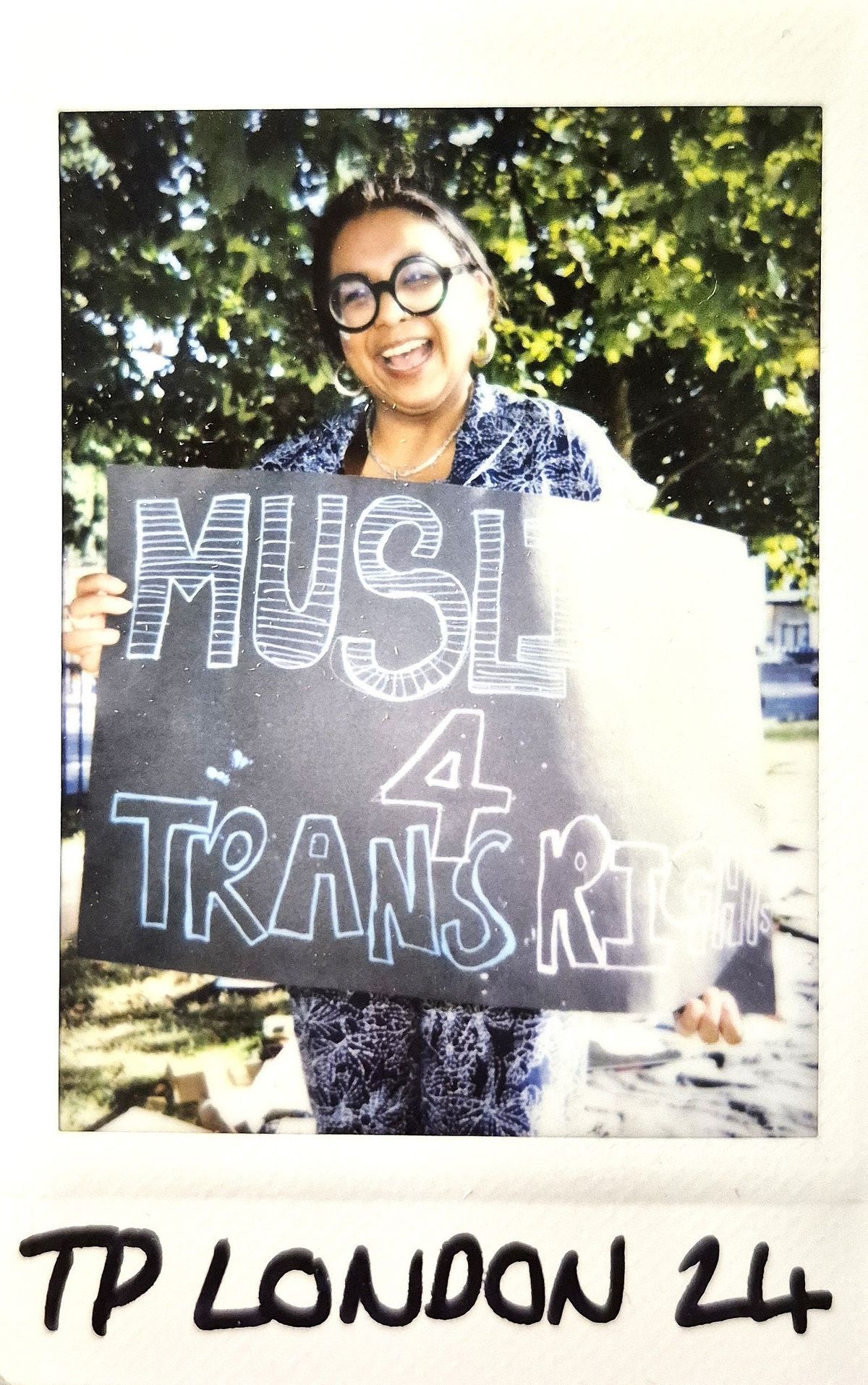 A smiling individual holds a sign that reads, "MUSLIMS 4 TRANS RIGHTS,"