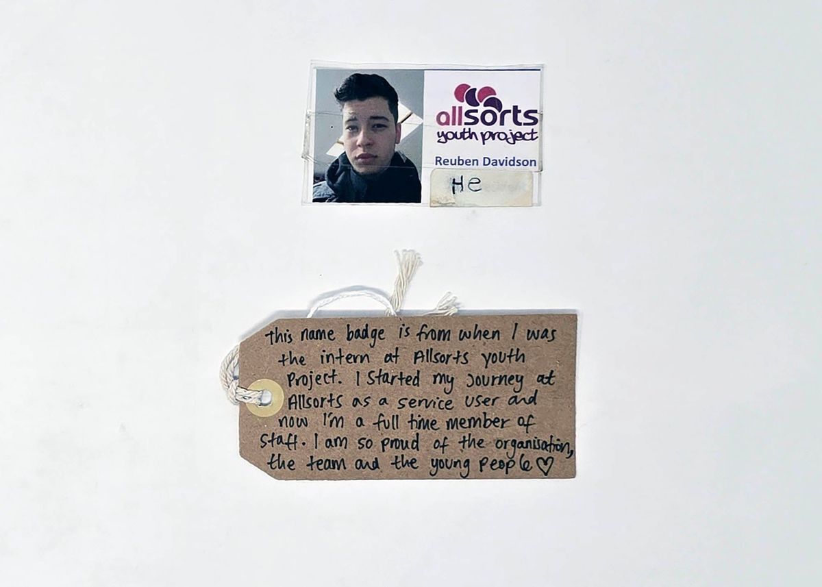 Name badge & tag; Allsorts Youth Project; name "Reuben Davidson" with photo wearing black scarf. Sticker handwritten "He". Card laminated, repaired in sellotape on back. Card creased across face in photo.