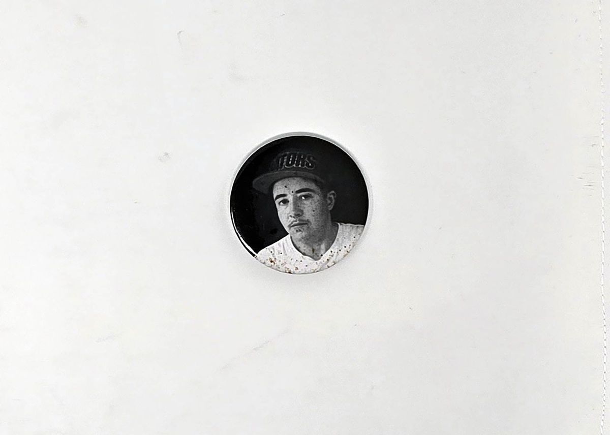 Small black badge showing a black and white portrait of Darcy wearing a white tshirt and black hat.