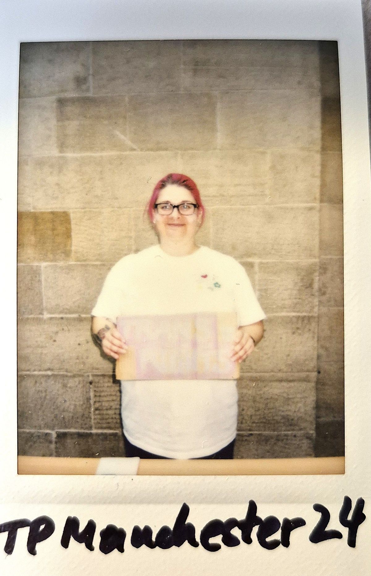 A person with pink hair and glasses holds a sign, standing against a textured stone wall background.