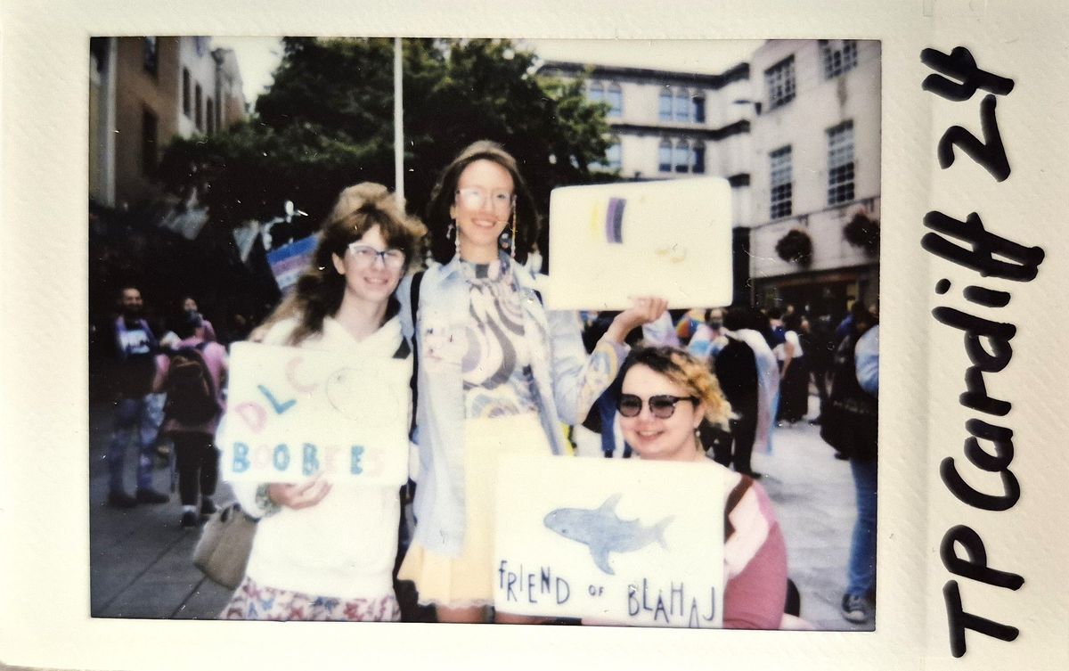 Three people are smiling and holding signs in a lively outdoor setting at a public event.