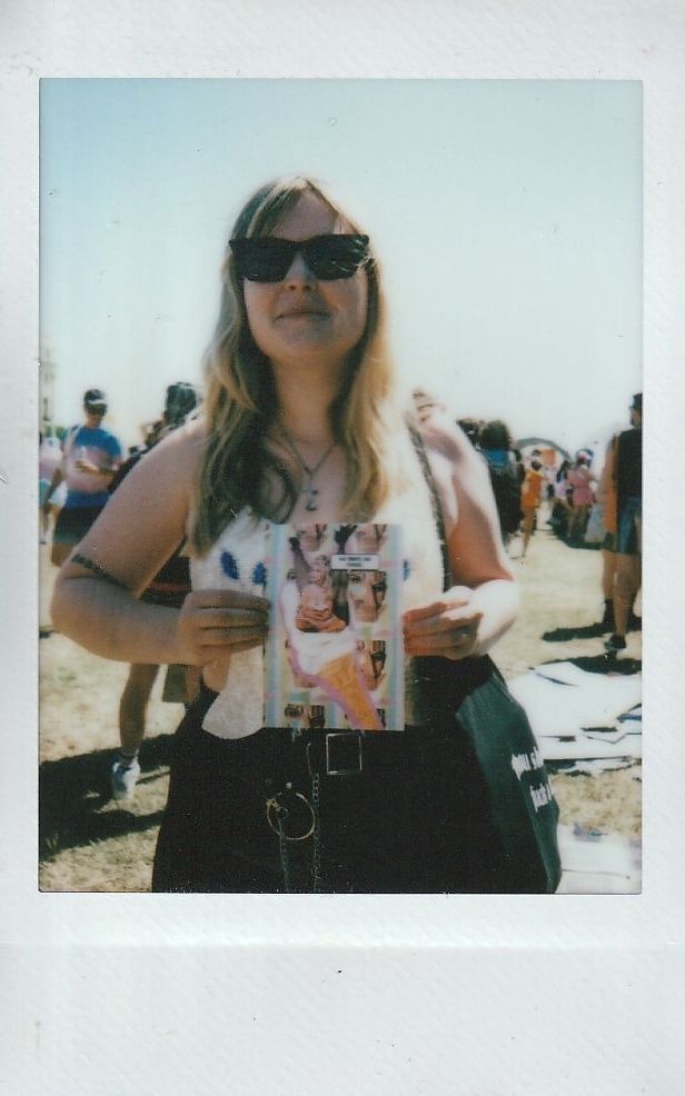 A woman wearing sunglasses holds a colorful magazine, standing outdoors among a crowd on a sunny day.