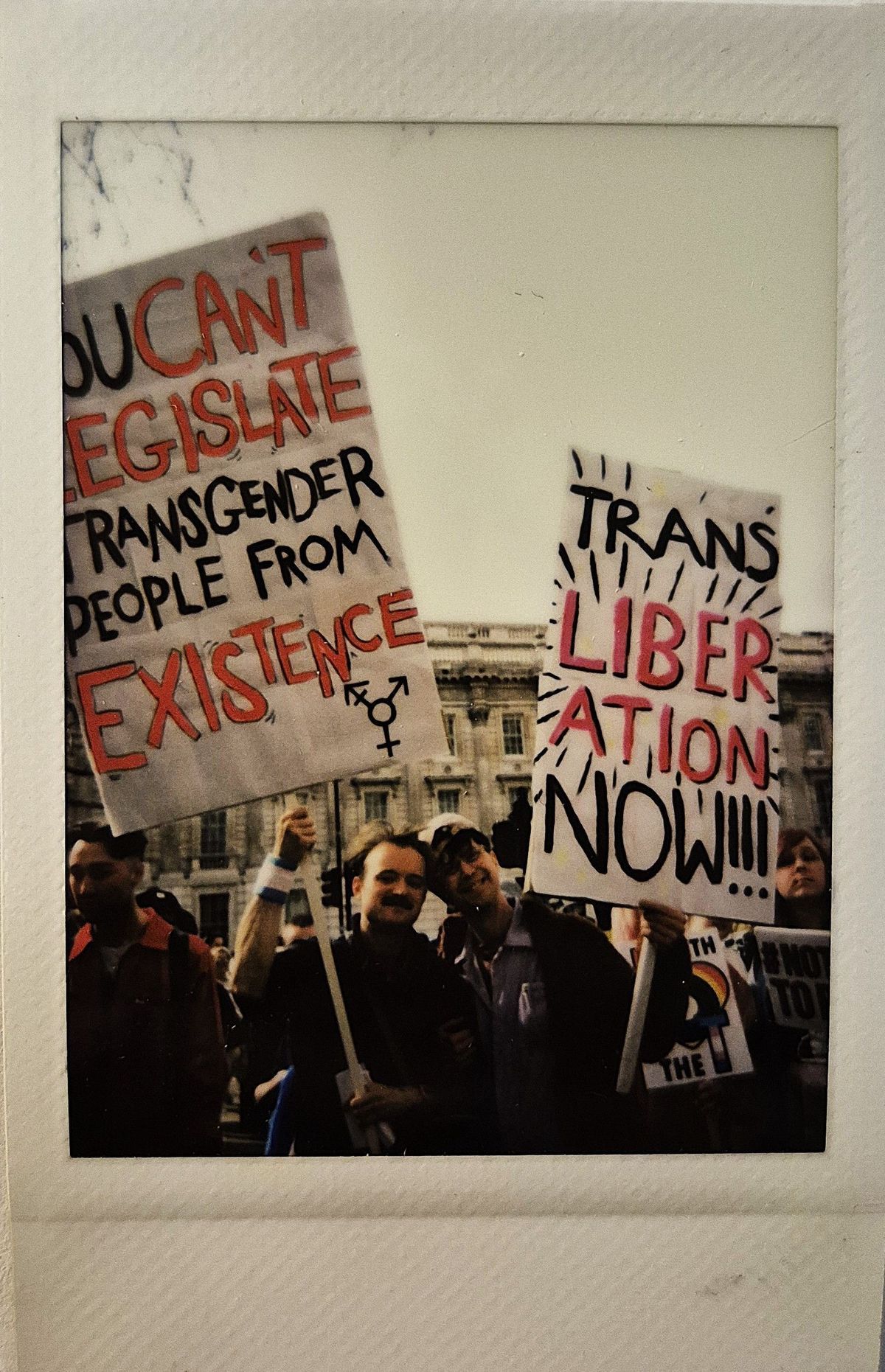 The image shows people holding signs advocating for transgender rights and liberation including one sign which says "TRANS LIBERATION NOW!!"