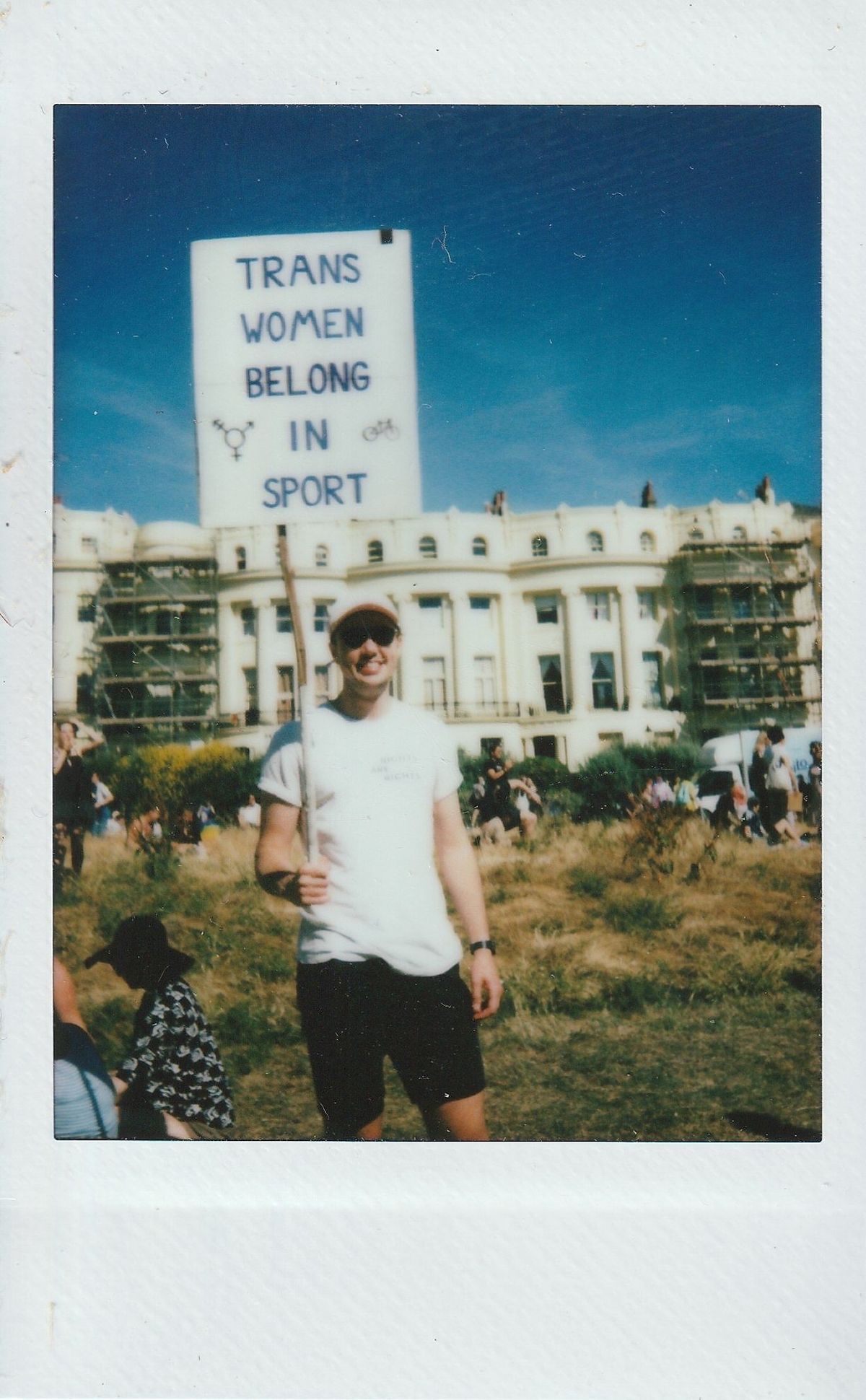 A person is holding a sign stating "Trans Women Belong in Sport" in an outdoor setting.