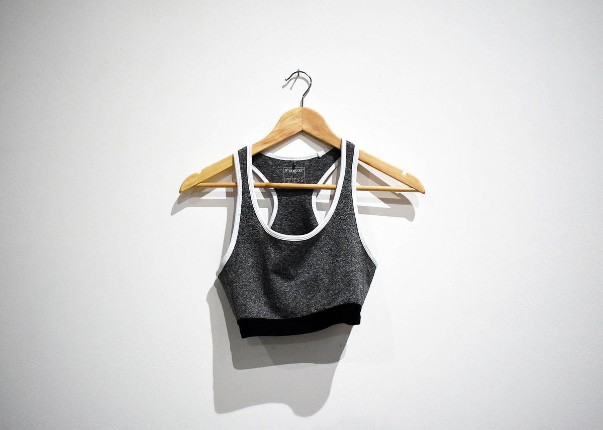 A gray sports bra with white trim hangs on a wooden hanger against a plain wall.