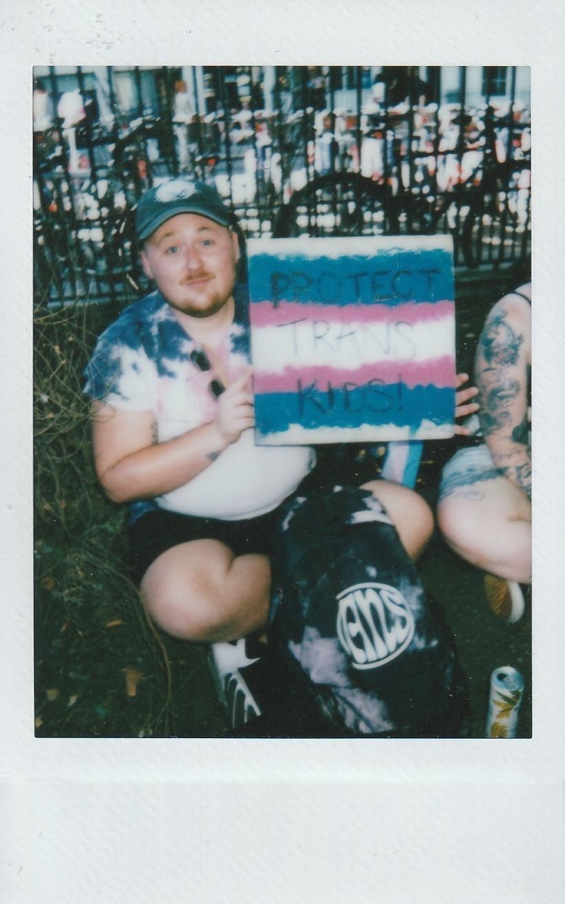 A person sits on the ground, holding a sign that reads "Protect Trans Kids" with colorful background