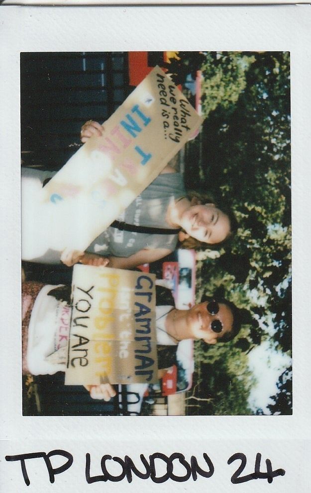 Two individuals hold signs with messages at an outdoor event, smiling and surrounded by trees.