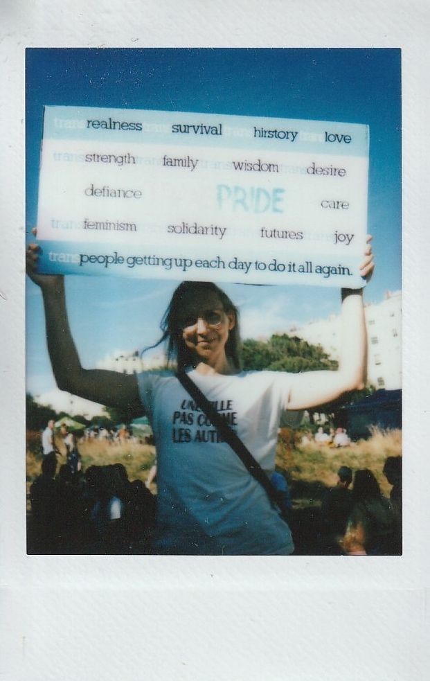 A person holds a sign with words like "realness" and "Pride," standing among a crowd of people.