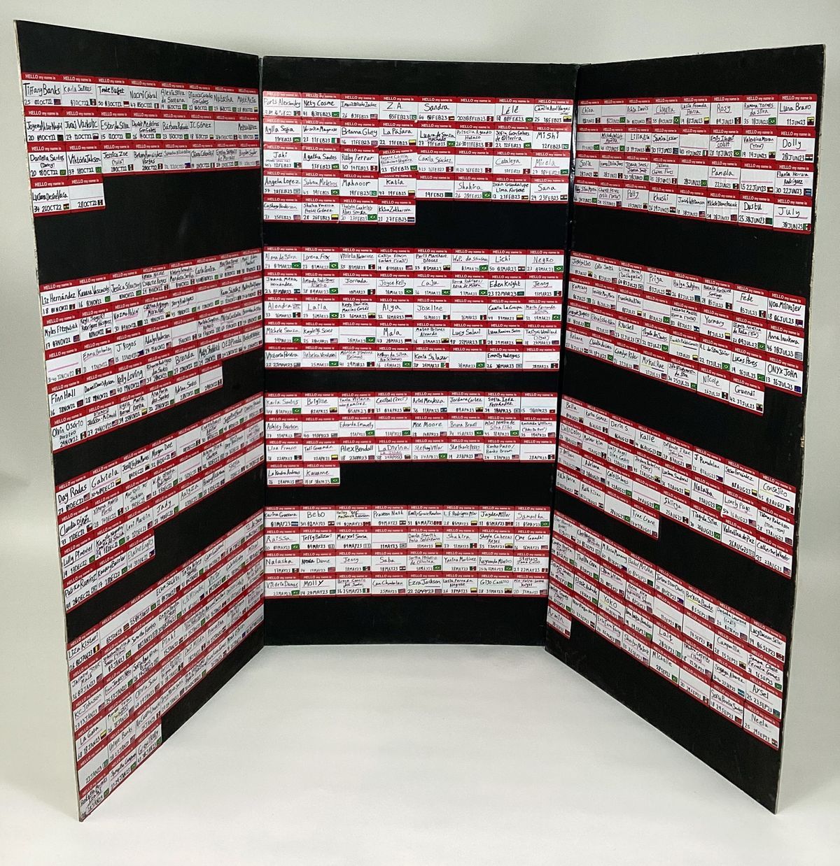 A mural for Transgender Day of Remembrance 2023 entitled "Hello, My Name Was ...". It is a three sided black display stand covered with 392 red and white name badges. Each name badge has a name from the TDoR Remembering Our Dead website.