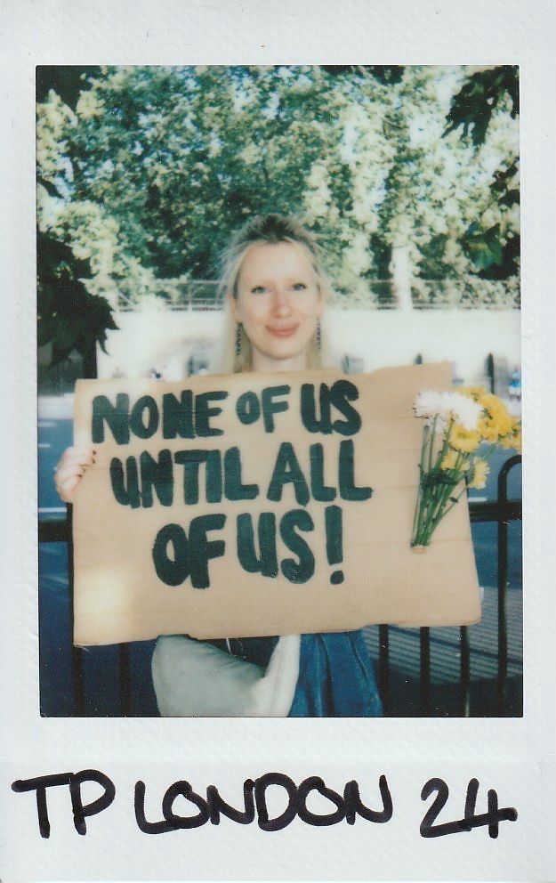 A person holds a cardboard sign reading "None of us until all of us!" with flowers attached.