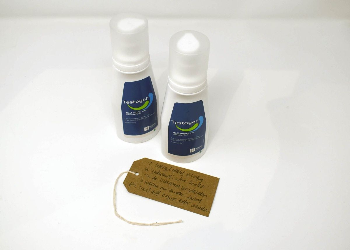 Two bottles of Testogel. The bottles are white and have a dark blue label with a green and light blue splash logo. With a brown, handwritten luggage tag.
