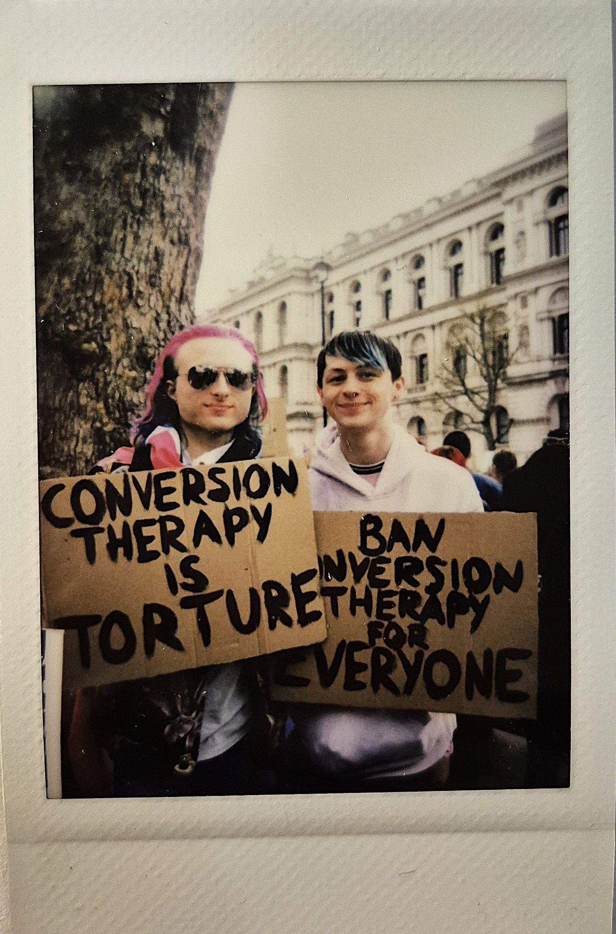 Two people hold protest signs which says "CONVERSION THERAPY IS TORTURE" and "BAN CONVERSION THERAPY FOR EVERYONE".