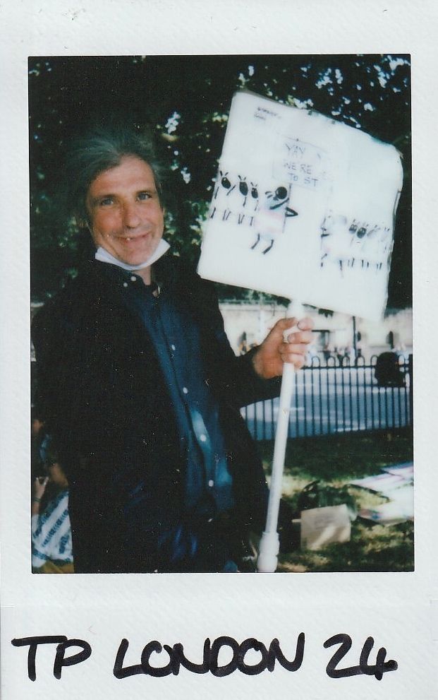 A person stands in a park holding a sign with illustrations depicting trans people.