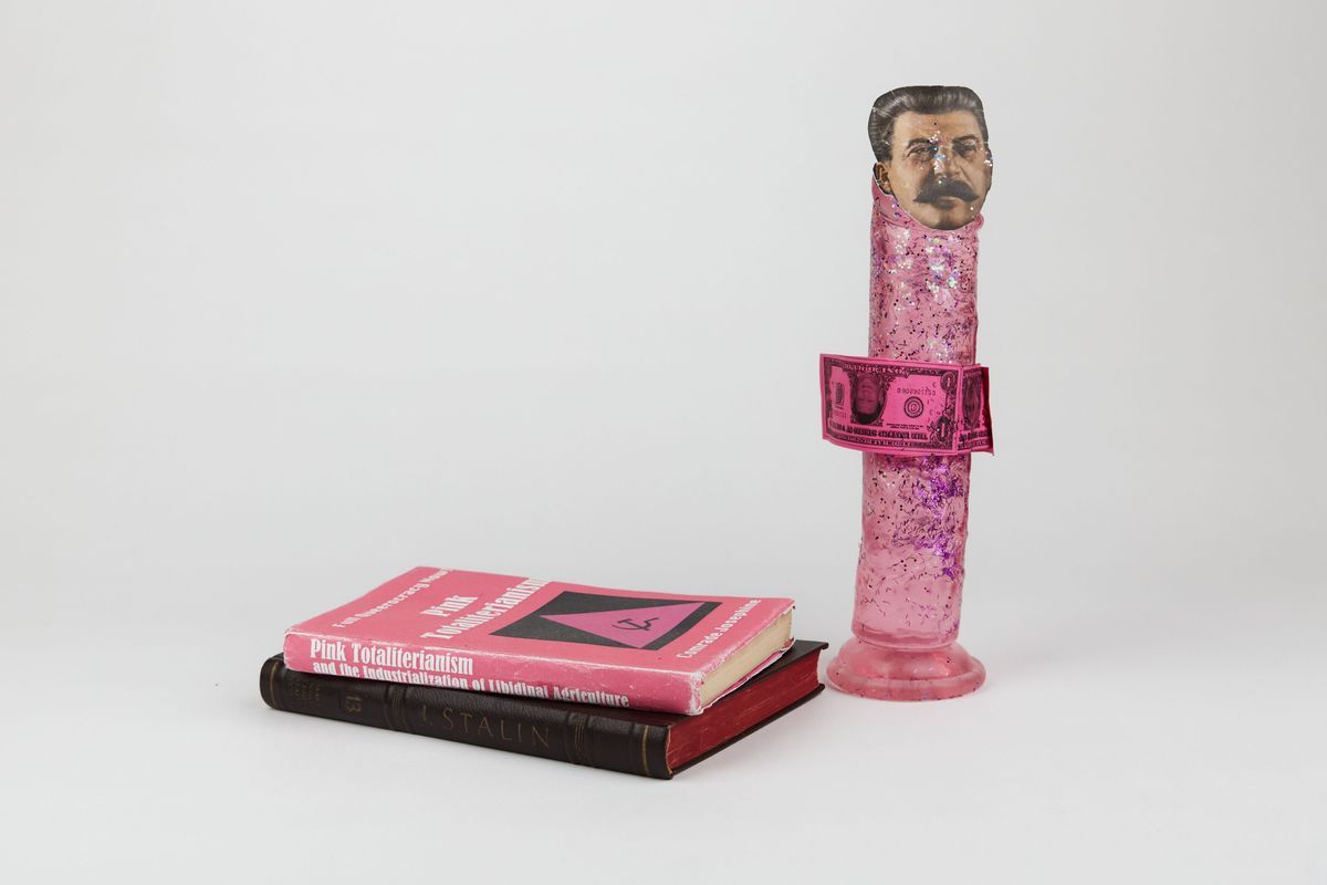 A pink, glitter-covered dildo with a a cutout of Stalin's face on the top stands beside two books stacked on top of each other. The top book is pink with the title "Pink Totaliterianism."