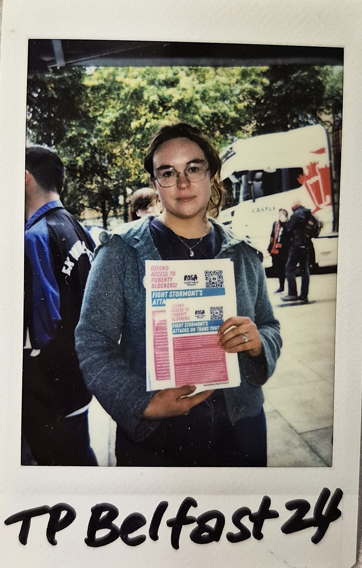 A person holding flyers stands outside, with trees and a bus in the background while wearing glasses.