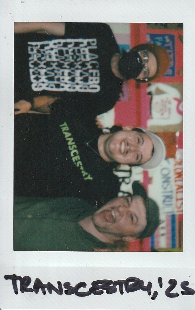 Three people are happily posing together, wearing casual clothes and hats, against a colorful background.