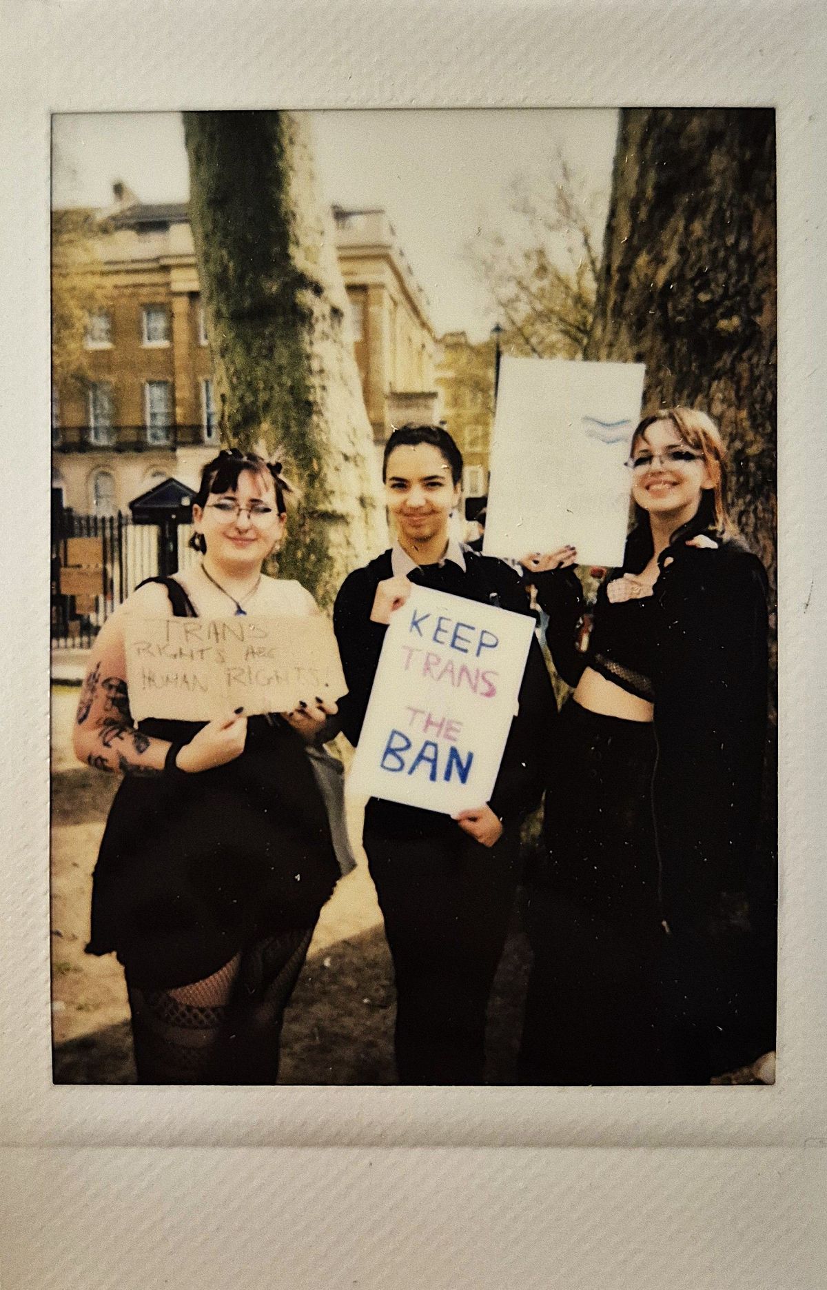 Three people are holding signs advocating for trans rights with phrases such as "KEEP TRANS IN THE BAN" beside large trees outdoors.