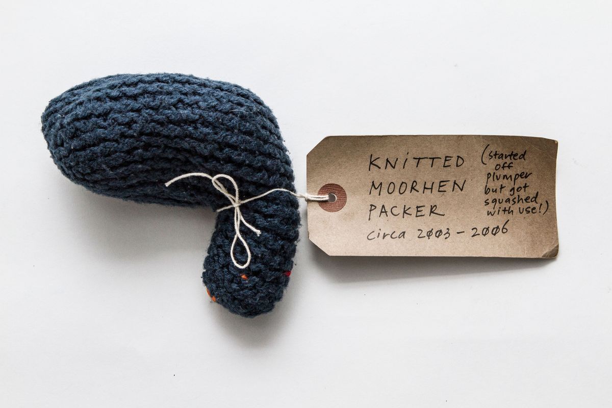 Dark blue knitted packer. a few bits of orange yarn poke through the blue stitches at the top tight of the packer. Thers is a white tag tied around the top.