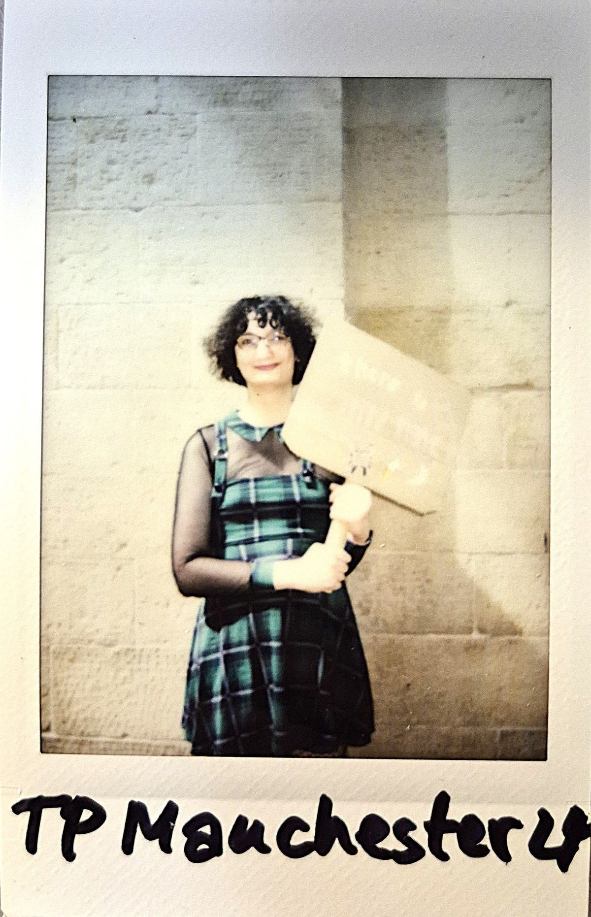 A person stands holding a sign, wearing glasses and a plaid dress, against a stone wall.
