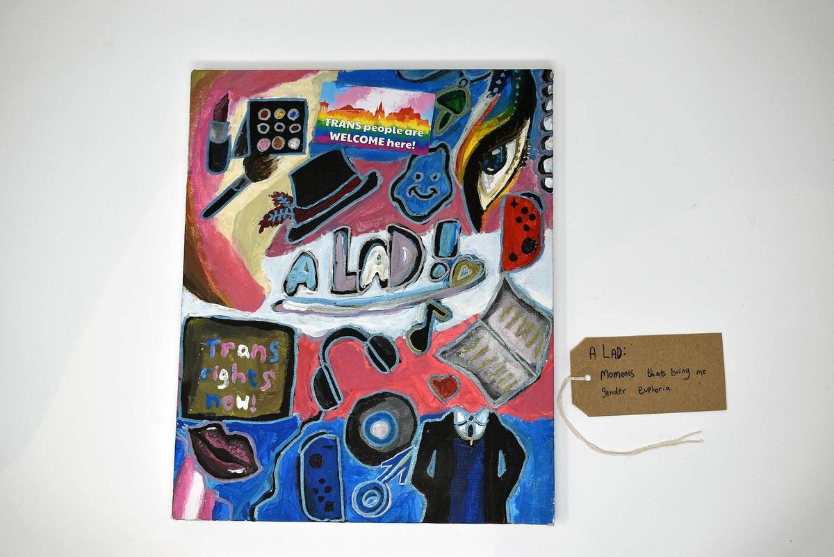 The colorful artwork features symbols and messages supporting transgender rights with the text "A LAD!" in the centre. With a brown handwritten luggage tag.