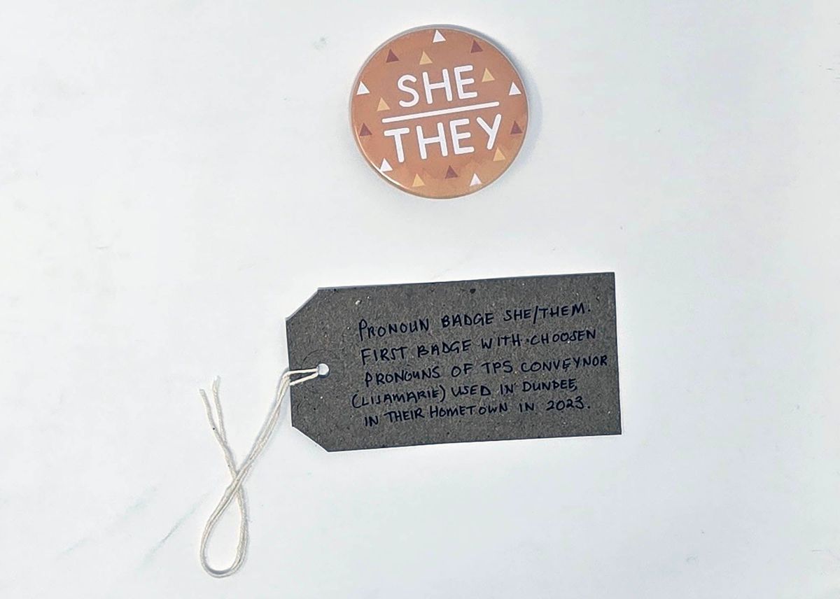 The image shows a button with "She/They" above a gray tag with handwritten notes.