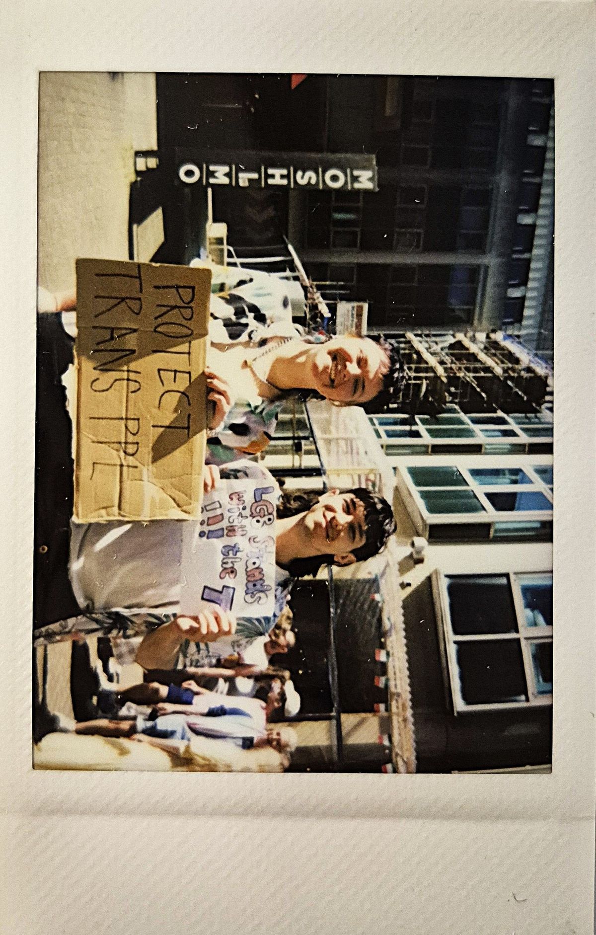 Two people are smiling at a gathering, holding signs which say "PROTECT TRANS PPL".