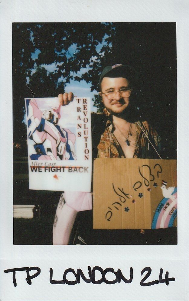 A person holds two signs expressing themes of trans pride and activism while smiling outdoors.
