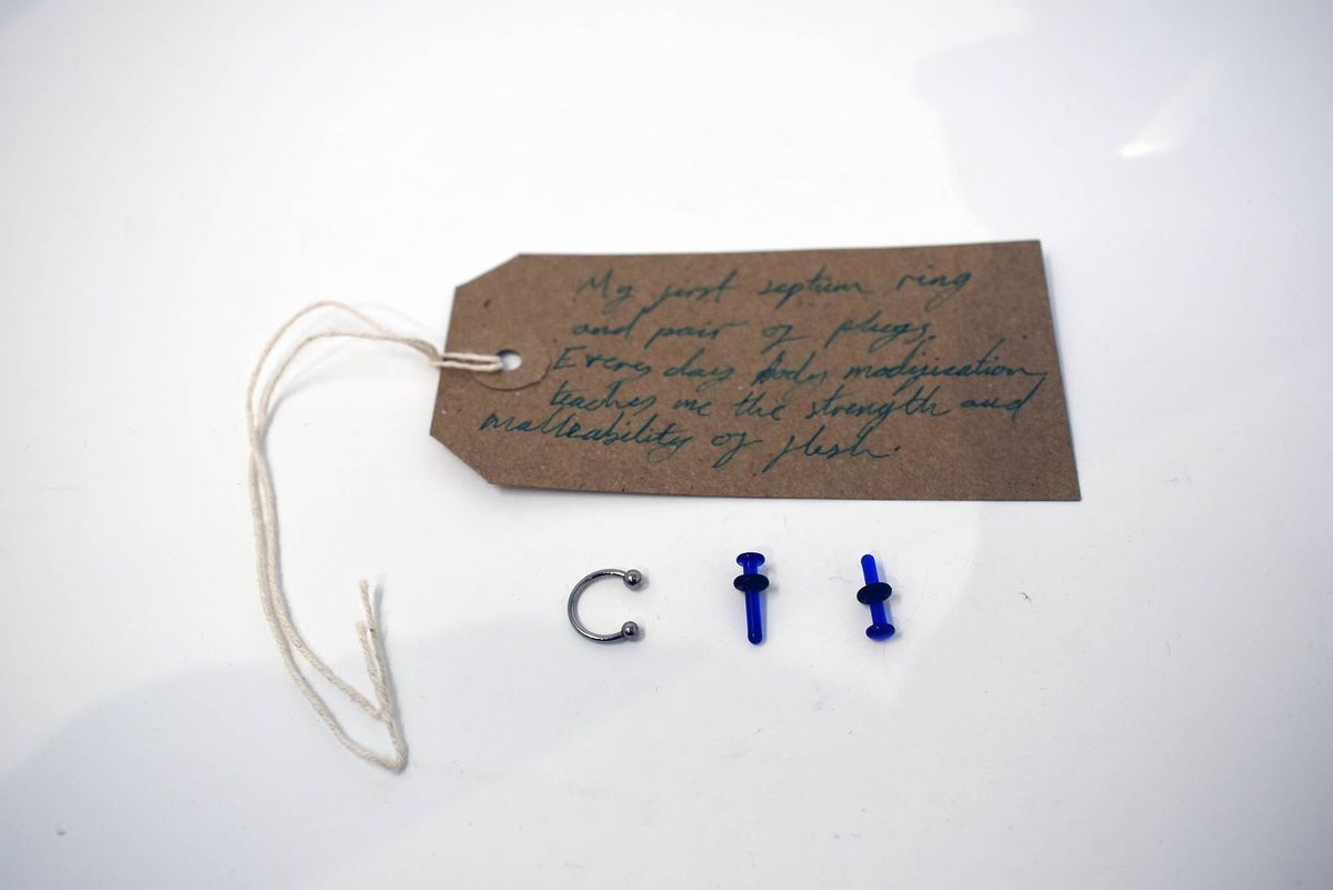 A handwritten tag, metal septum ring, and blue plugs are displayed on a white surface.