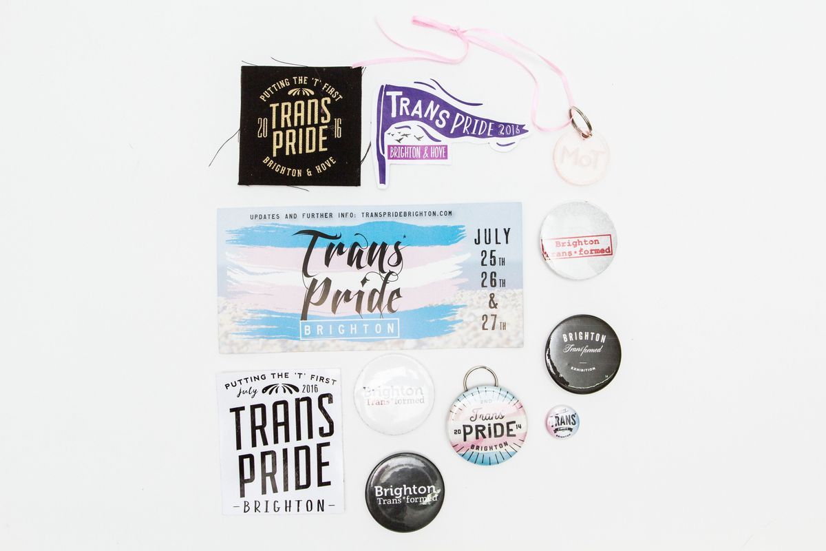 The image displays a collection of memorabilia related to Trans Pride events in Brighton, including various badges and a ticket stub. The items feature phrases such as "Putting the 'T' First" and "Trans Pride Brighton & Hove," with some representing specific years like 2013 and 2014. The colorful designs incorporate the colors pink, blue, black, and white, highlighting the spirit of the LGBTQ+ community in Brighton. The main ticket prominently showcases the dates July 25th, 26.