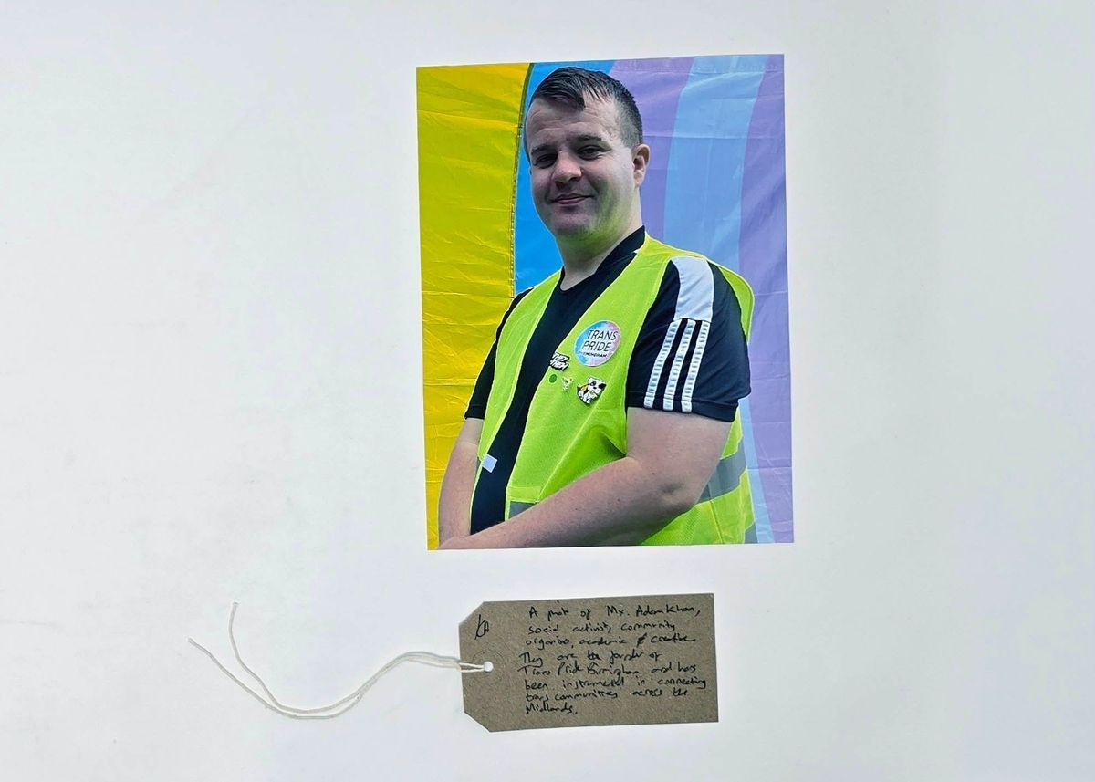 A person wearing a yellow vest with "Trans Pride" stands against a colorful background and a handwritten note