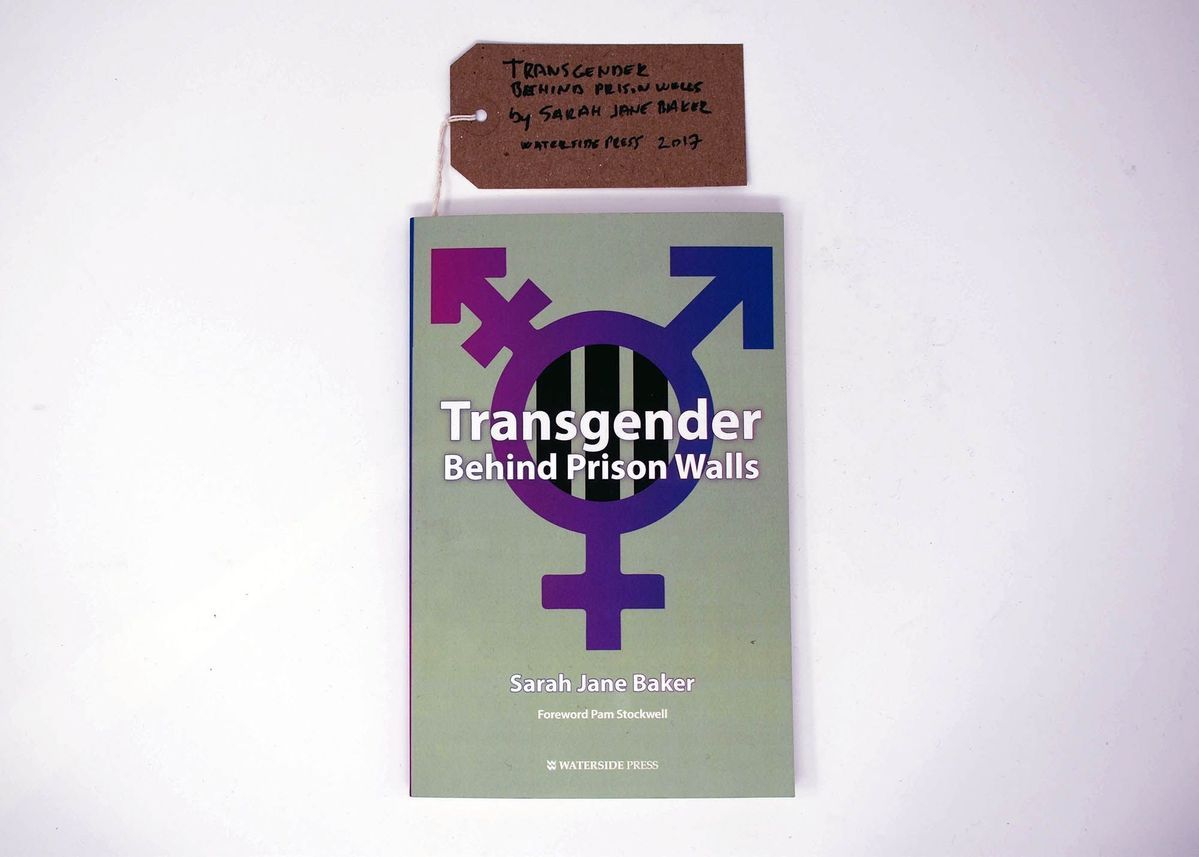 A book called 'Transgender Behind Prison Walls' by Sarah Jane Baker which has a grey background and the transgender symbol in a gradient pink and purple hue.
