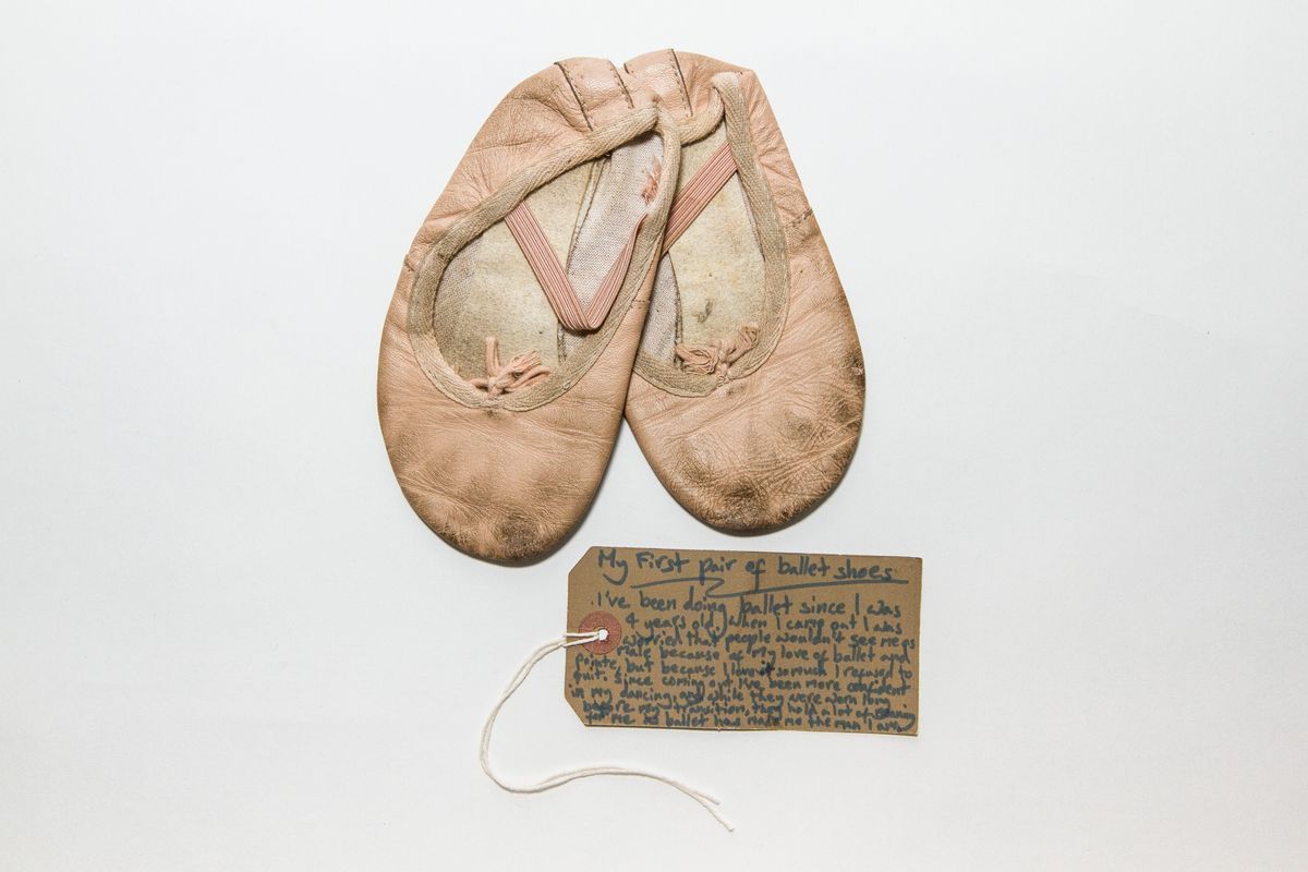 Pink ballet shoes tied together with a small piece of white string. Both shoes have black marks on the bottom and show other signs of wear such as dirty elastic over each bridge and frayed seams.