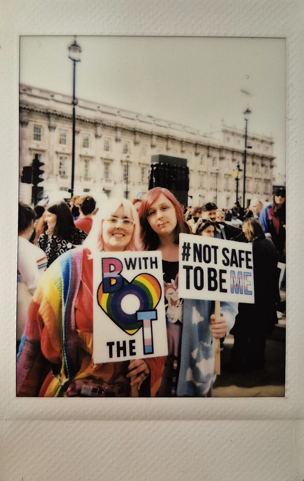 Two people holding colorful protest signs which say "B WITH THE T" and "#NOT SAFE TO BE ME".