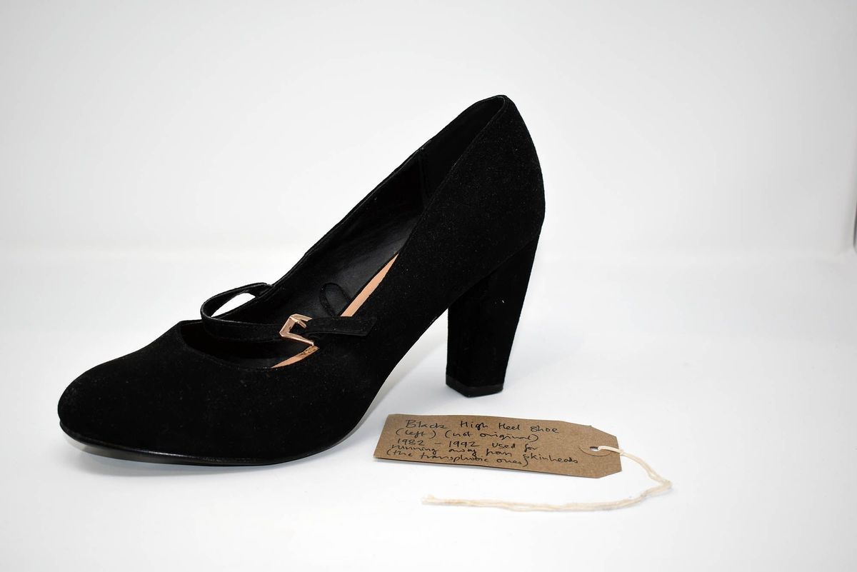 The image shows a black high-heeled shoe with a strap, accompanied by a handwritten tag.