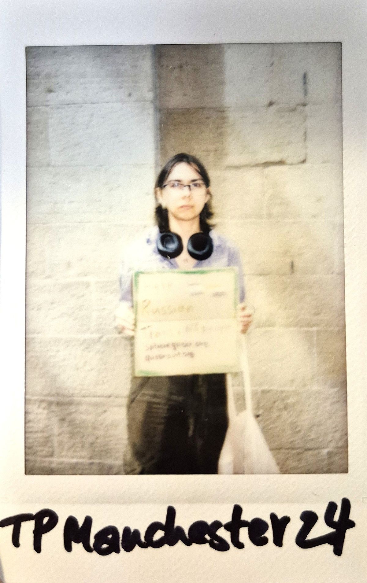 A person standing against a brick wall holding a handwritten sign, with headphones around their neck.