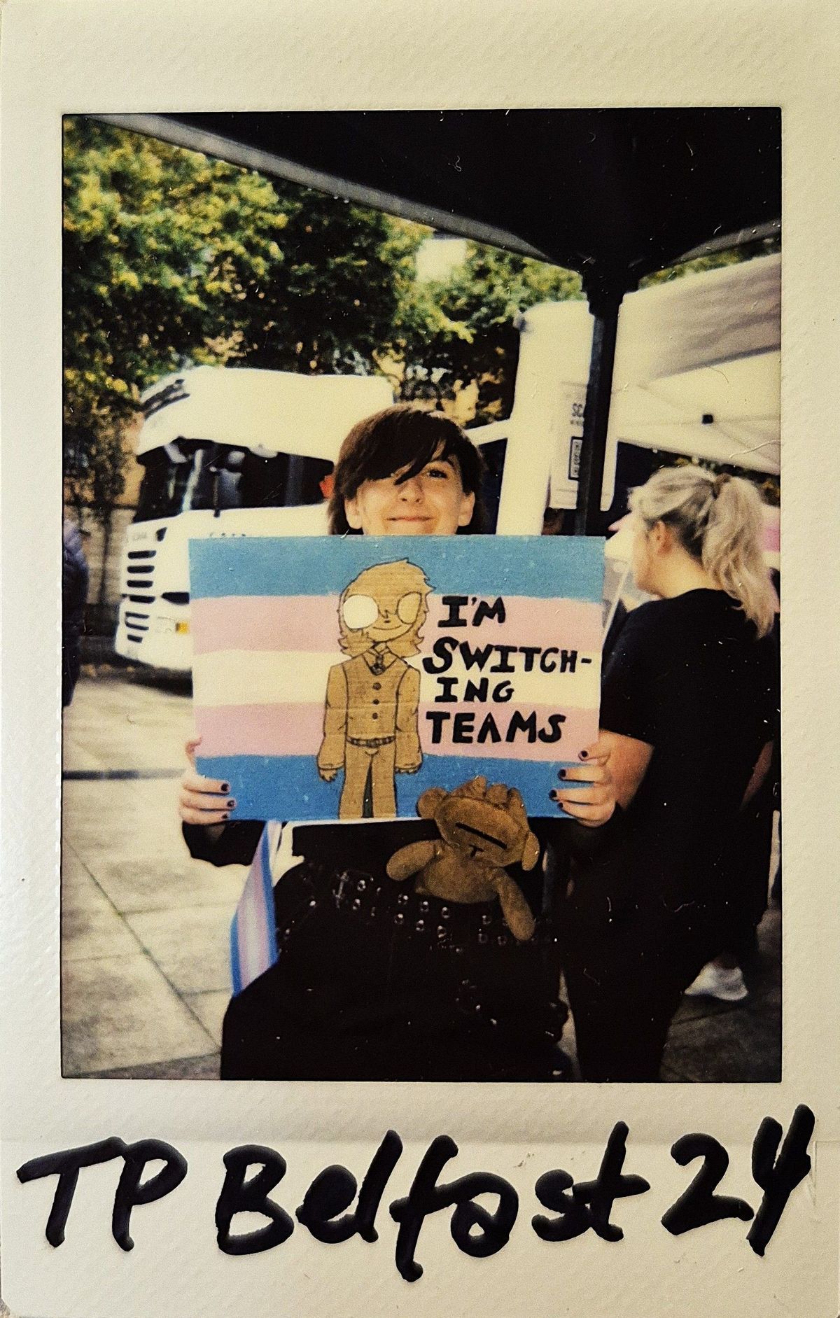 A person is smiling while holding a sign with a drawing and the text "I'M SWITCHING TEAMS".