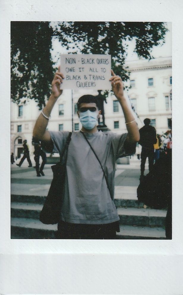 A person wearing a mask holds a sign which says "Non-Black Queers Owe It All To Black & Trans Queers".