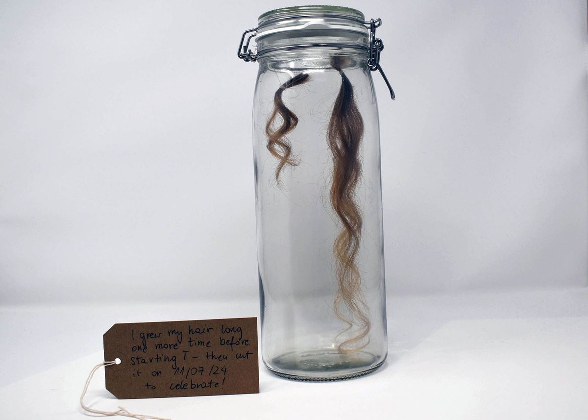A clear glass Mason Jar containing two locks of brown, human hair, suspended on thread. One lock is longer than the other. With a brown, handwritten luggage tag.