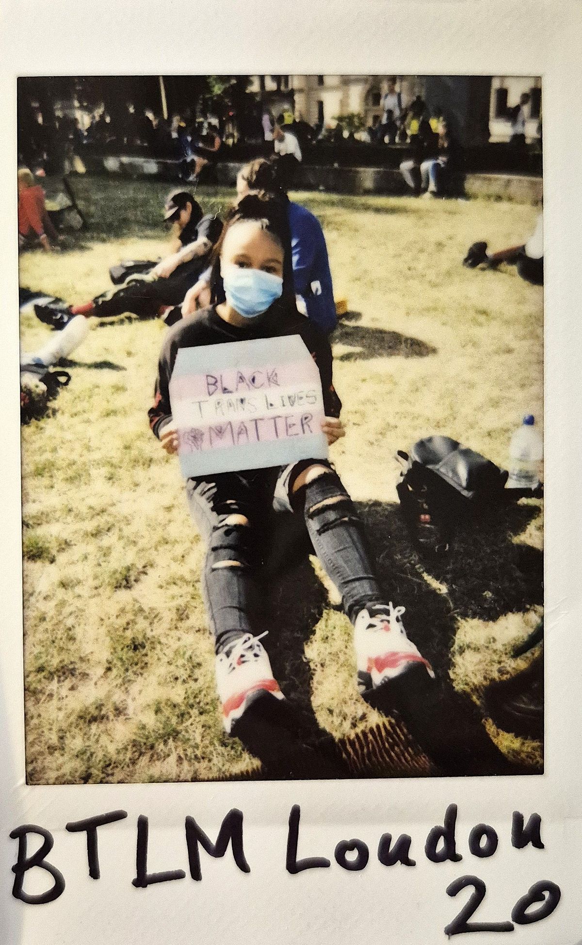 A person sits outdoors, wearing a mask, holding a sign supporting Black trans lives.