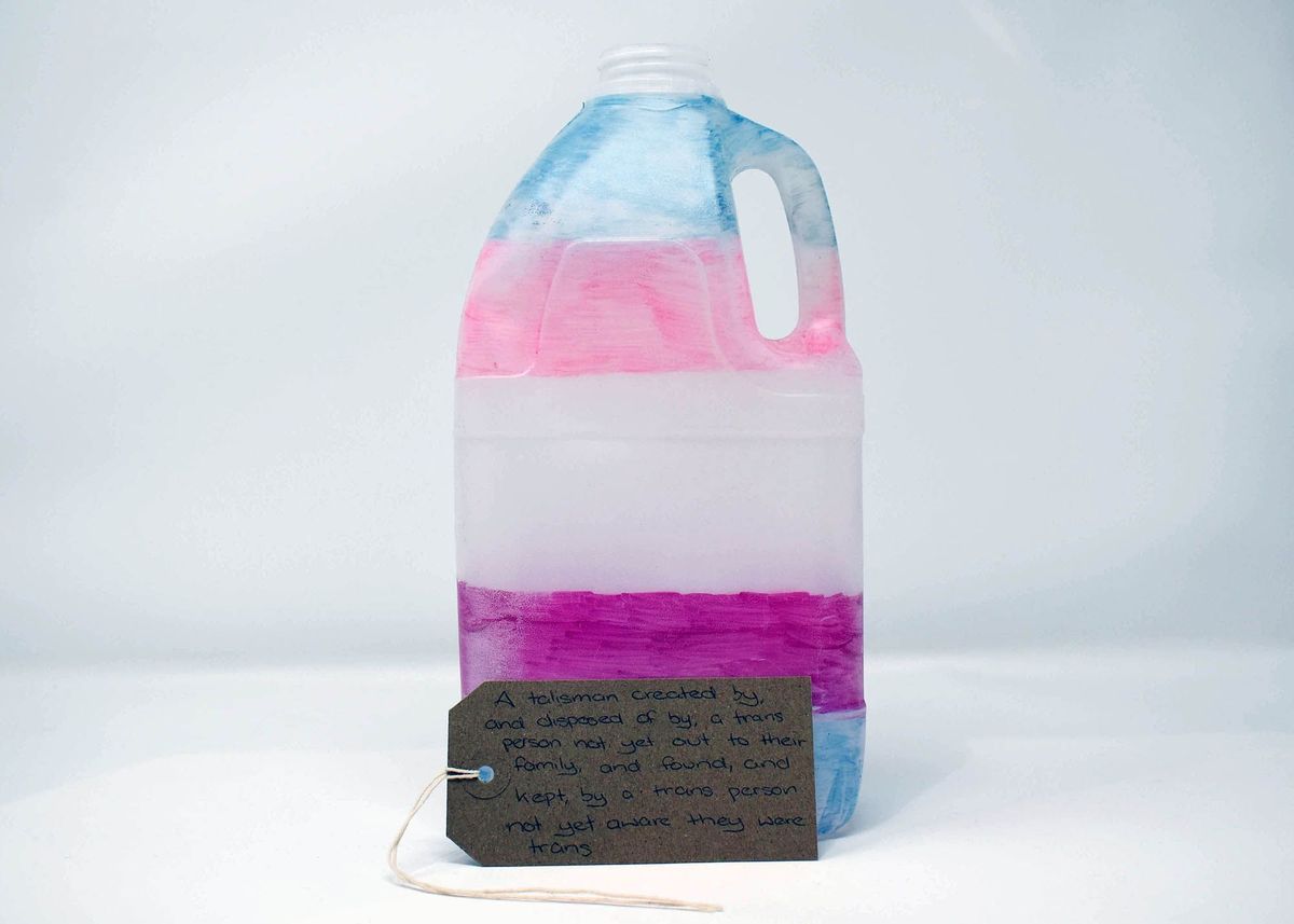 A plastic bottle hand coloured in the trans flag colours, with a brown luggage tag