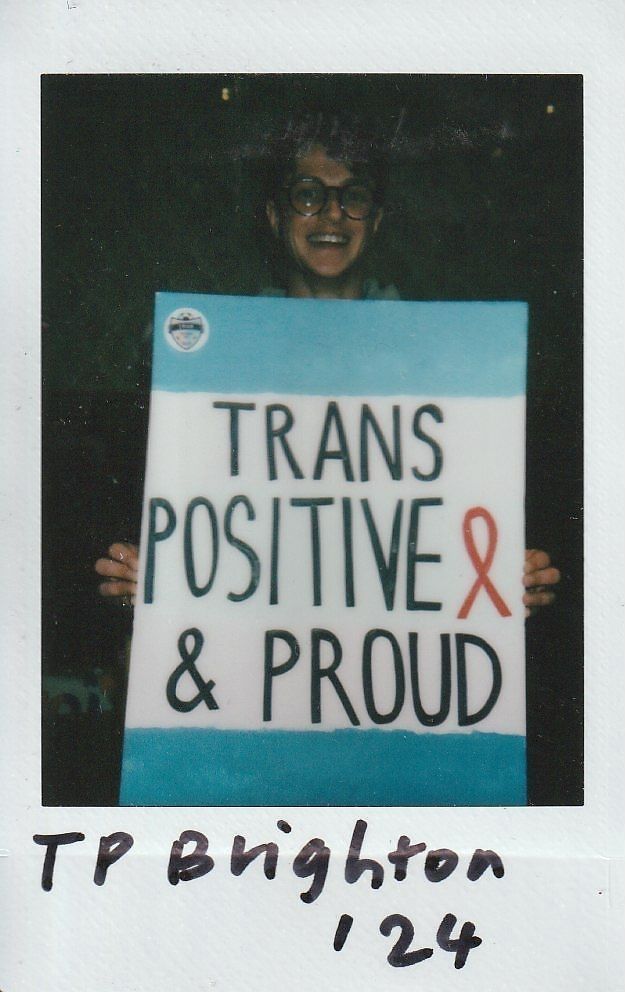 A person is smiling while holding a sign that says, "Trans Positive & Proud," in bold text.