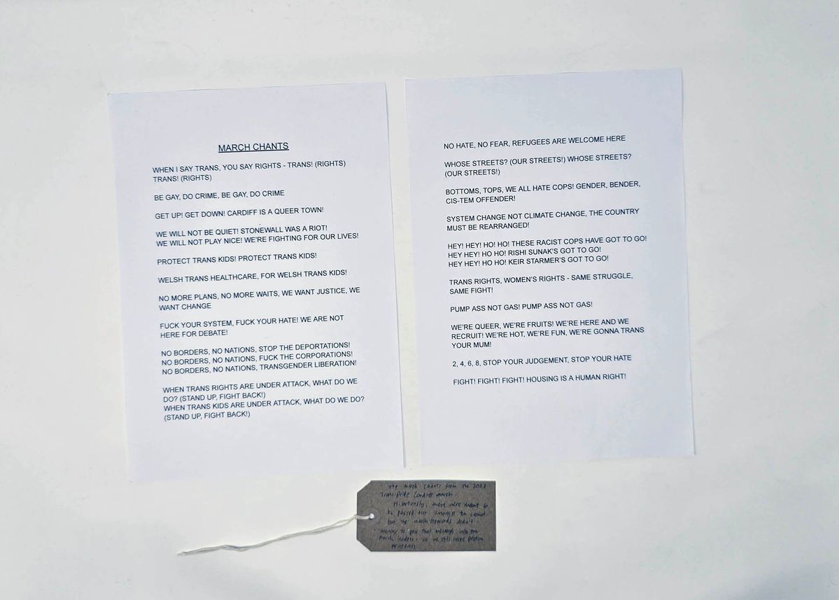 Two sheets of white A4 paper printed in black text with a list of chants from the 2023 Trans Pride Cardiff march. With a brown handwritten luggage tag.