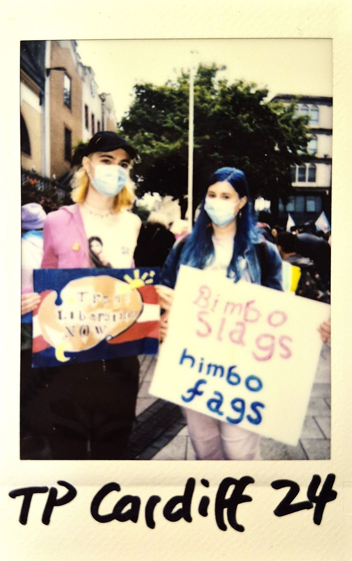 Two people with masks hold colorful signs at a public gathering.