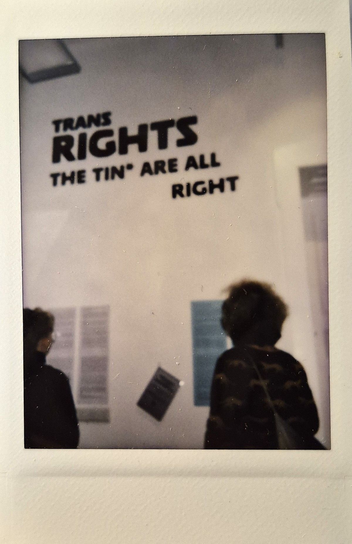 The image shows two people looking at a wall with a sign reading "Trans Rights The Tin* Are All Right".
