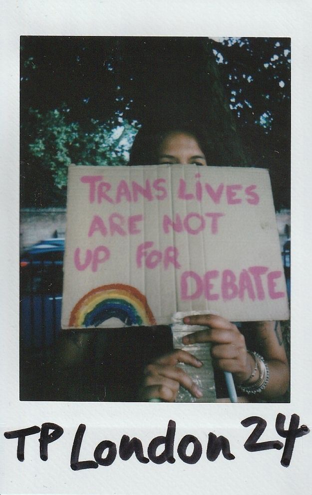 A person holds a cardboard sign stating, "Trans lives are not up for debate," featuring a rainbow.