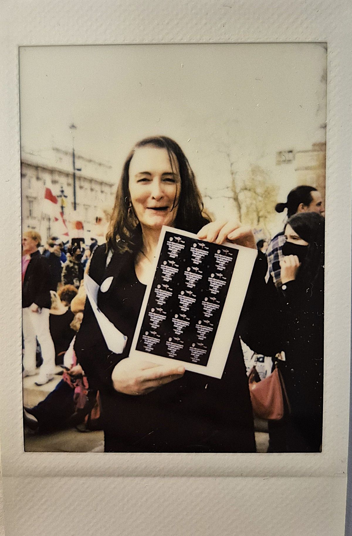 A person with long hair is smiling, holding a document in a busy outdoor setting.