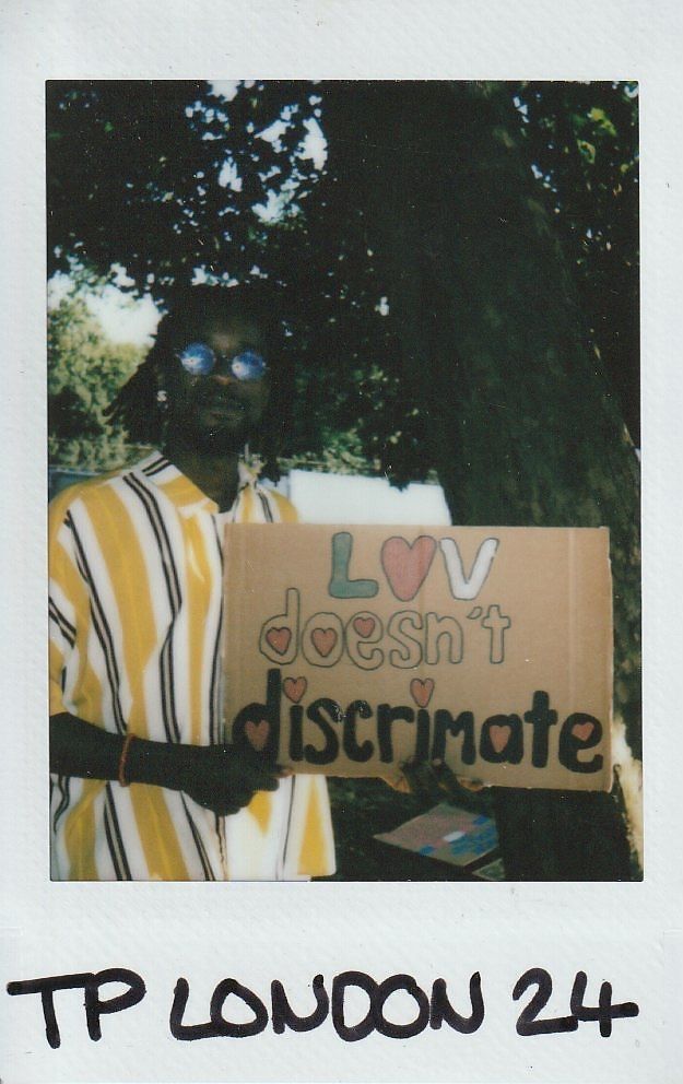 A person stands holding a cardboard sign that says, "Love doesn't discriminate," under a tree.
